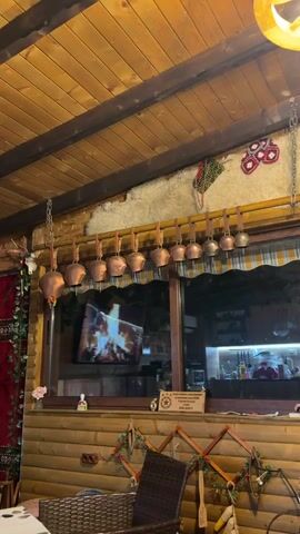 The camera pans across the rustic interior of a restaurant or dining area. The ceiling is lined with wooden beams, from which a row of cowbells hangs. Below, a window displays a television screen showing a fire. The walls are clad in wood panels and decorated with traditional textiles and folk art. Tables with wicker chairs are visible, and a boy is seen sitting at one table, using a tablet. The overall ambiance is warm and traditional, with a touch of local culture reflected in the décor.