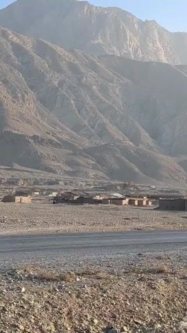 The video shows a landscape featuring a large mountain range in the background under a clear blue sky. The ground is arid and rocky. A straight paved road runs across the middle ground. On the side of the road closer to the mountains, there are scattered, modest, one-story buildings that appear to form a small village. A dark-colored SUV is seen driving on the road. The video appears to be taken from the side of the road.