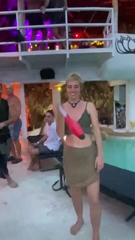 The video shows a woman in a green crop top and short brown skirt juggling a red juggling club. She is at a party next to a pool with other people around. There is a man in a black outfit talking to her. Another person is sitting on a chair behind them, and several people are on a second-story balcony above them. The building is decorated with lights.
