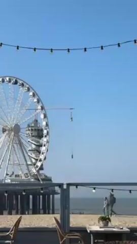 The video shows a seaside scene with a large Ferris wheel to the left of a pier extending into the sea. A construction crane is visible behind the Ferris wheel. A person is bungee jumping from the crane, falling towards the ground. In the foreground, there's a beachside cafe or restaurant setting with chairs, tables, and string lights overhead. Two people are walking along the beach near the water's edge. The sky is clear and blue, indicating a sunny day.