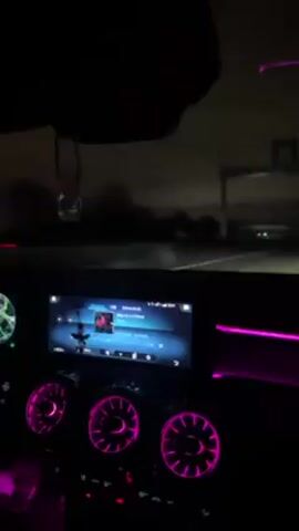 The video shows the interior of a car driving at night, focusing on the dashboard. The car's infotainment screen displays music, and the air vents are illuminated with pink light. The car is driving on a highway, visible through the windshield. The environment is dark, with occasional streetlights and the headlights of other cars. The scene has a modern, stylish aesthetic due to the car's interior design and the ambient lighting.