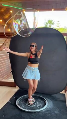 A woman stands in a room with wood-paneled walls and an outdoor background visible through a window. She is wearing sunglasses, a black crop top, and a denim skirt. She is surrounded by large soap bubbles. In the foreground, she is standing within a circular metal frame on a black mat. She is interacting with the soap bubbles, at one point appearing to take a bite out of one. She then laughs and covers her mouth. A black screen is behind her.