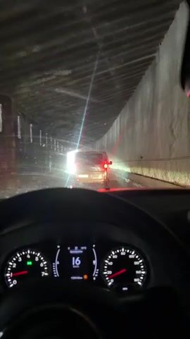 A driver's perspective inside a car moving through a tunnel is captured. The speedometer reads around 17 km/h. The tunnel has what appear to be concrete walls and a low ceiling. The driver follows closely behind a van, its brake lights visible. The tunnel appears damp with some water on the road. The end of the tunnel is visible in the distance, brightly illuminated.
