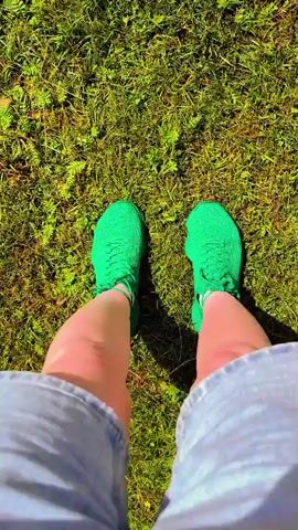 The video begins with a close-up shot of a person's legs wearing bright green athletic shoes, standing on a patch of green grass. The camera then pans down to show a pair of blue swim fins lying on the grass. The scene transitions to a wider shot of a lake surrounded by trees, with a grassy area in the foreground and a cloudy sky above. The last frame shows the swim fins again, with the same lake and sky in the background. The video appears to be a short promotional clip for a swimming or water activity.