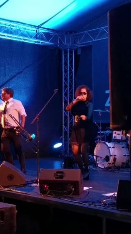 On a stage bathed in blue light, a band is performing. To the left, a man in a white shirt, black tie, and dark pants holds a trombone. Another man joins him, also with a trombone, and they stand near the front. The lead singer, a woman with curly hair and sunglasses, is positioned center stage, singing into a microphone. She wears a dark dress with a lace hem and black stockings. Various speakers and monitors sit along the front edge of the stage. A drum set is visible behind the performers, near the back of the stage. The backdrop consists of a dark wall, likely a curtain, while the stage is covered by a temporary frame structure, indicating an outdoor event or festival setup.