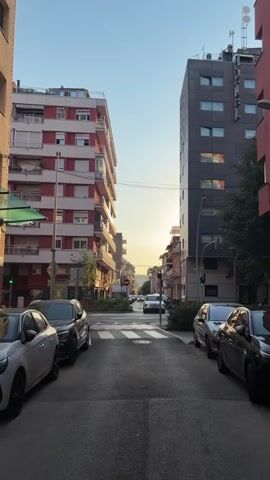 The video shows a street view in a city. On the left side of the street, there is a tall, multi-story building with a red and white facade, featuring balconies. On the right side, there is a darker, taller building. Several cars are parked along both sides of the street. A white car is crossing a pedestrian crossing in the middle of the street. In the distance, more buildings and trees are visible, and the sky is clear with the sun setting or rising, casting a warm light. The overall environment appears to be an urban residential area. No people are clearly visible or interacting in the frame.