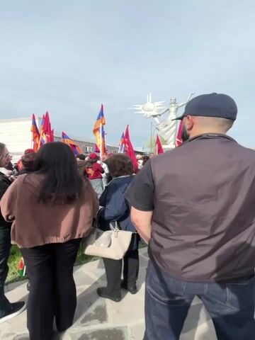 The video shows a crowd of people gathered around a monument in Armenia. There are many Armenian flags being waved. Most of the people are standing and looking towards the monument. Some are holding phones, possibly taking pictures or videos. The monument appears to be a statue of a woman holding a sun emblem. The sky is overcast, and the setting seems to be a public square or park.