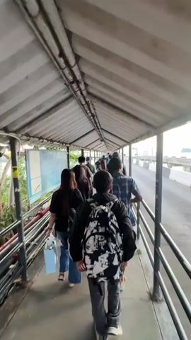 The video shows a person walking down a covered walkway towards a pier or bridge. There are other people walking in the same direction. The walkway has a corrugated metal roof and a railing on the right side. Beyond the railing is a road or pathway with vehicles. In the distance, there is the ocean and a small island. The sky is cloudy. The video pans slightly to the right, revealing more of the pier and the ocean. There are streetlights along the pier. The video zooms out slightly at the end, showing the scene from a wider perspective.