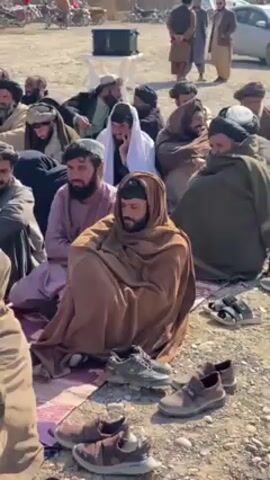 The video shows a group of men sitting on the ground in an outdoor area. They are all dressed in traditional clothing and appear to be listening to someone speaking. Many of them are wearing head coverings and are wrapped in blankets or shawls. There are cars and motorcycles parked in the background, along with some dry vegetation. The ground is dusty and rocky.