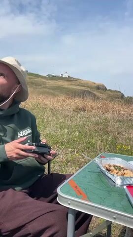 A video shows two men having a picnic on a green, grassy hillside overlooking the sea. A white lighthouse is visible in the distance. The first man is operating a drone remote, while the second man sits at a small green folding table with a white wine bottle, a container of grapes and sliced bananas, and a foil container of food. The man at the table is wearing dark sunglasses and looks directly at the camera. The sky is mostly blue with some clouds.