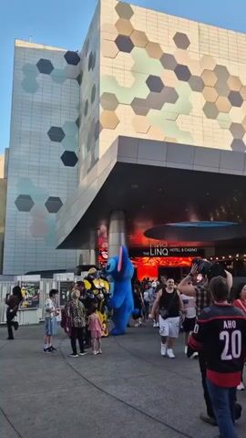 The video shows a bustling outdoor area in Las Vegas, Nevada, at dusk. People are seen walking past The LINQ Hotel & Casino, a building with a distinctive honeycomb facade. The area is crowded, with people of diverse ages and ethnicities. Some are holding hands, while others are dressed in casual attire. A person in a Stitch costume interacts with the crowd. In the background, palm trees and other buildings are visible, including a tall LED sign advertising "Spiegelworld.com". The overall scene suggests a vibrant and lively atmosphere.
