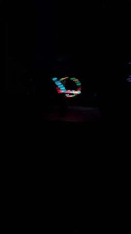 In a dark outdoor setting, a person is seen performing a light show with three light-up hula hoops. The hula hoops emit red, green, and blue lights. The person appears to be spinning and manipulating the hoops around their body. The surrounding area is dimly lit with a few distant lights visible. A sound resembling dance music plays in the background.