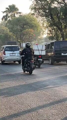 A group of motorcycles are traveling down a paved road. The road appears to be in a city or suburban area, with trees and buildings visible on either side. The sky is clear and the sun is shining, casting long shadows across the road. The motorcycles are of various makes and models, and some of them are carrying passengers or cargo. The riders are wearing helmets and casual clothing. The overall impression is one of daily life and transportation in a tropical or subtropical region.