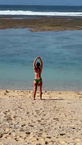 A woman in a green bikini is performing a stretching exercise on a sandy beach. She stands with her arms raised and clasped above her head, then bends forward at the waist, touching her hands to the sand. The ocean is in the background with waves and a rocky shore. The sun is shining.
