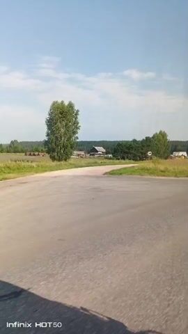 The video shows a scenic view of a rural landscape from a moving vehicle. The foreground is dominated by a paved road with a white dividing line. Beyond the road, there is a grassy field dotted with wildflowers and some dry grass. In the middle ground, there are several houses with reddish roofs and trees, suggesting a small village or settlement. The background consists of more trees and a vast, clear blue sky with a few scattered clouds. There are also traffic signs visible along the road. The lighting indicates it is daytime, likely a sunny afternoon. The camera appears to be mounted on a dashboard or is handheld by a passenger, providing a perspective from inside a moving vehicle. The overall impression is one of a peaceful, natural environment.