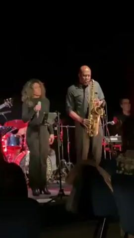 A musical performance takes place on a stage with a woman singing, accompanied by a saxophone player, a keyboardist, a drummer, and another musician who is only partially visible. The singer stands behind a music stand and holds a microphone, while the saxophonist plays. The keyboardist plays while looking at the sheet music and the drummer sits behind the drum set. The stage is dimly lit, with red lighting highlighting the drum kit. The audience, who are visible at the bottom of the frame, is watching the performance, with some members clapping.
