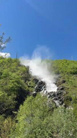The video shows a waterfall cascading down a rocky, green hillside under a clear, blue sky. The waterfall is powerful, creating a misty cloud as it hits the rocks. The surrounding area is lush with green trees and vegetation. In the distance, there are power lines silhouetted against the sky.