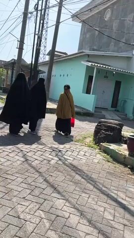 Three individuals wearing dark coverings and one individual wearing a yellow headscarf walk away from the camera down a paved alleyway with cobblestone-like pavers. Overhead power lines and utility poles are visible, as are houses with light blue and gray siding. The sun casts long shadows.