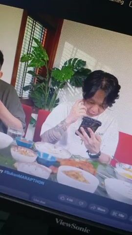 Here is a concise description of the video:

The video shows two men seated at a table laden with food. The man on the right is holding and looking at his phone, while the man on the left is eating.  The table is covered with a green banana leaf-like runner, and several bowls of food are arranged across it. Behind them, there's a window with blinds and a potted plant. The screen display indicates a live chat, suggesting the scene is part of a live streaming event, labelled "#BukanMARAPTHON."  The video is being viewed on a ViewSonic monitor.