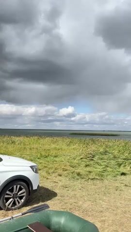 A static shot shows a grassy field with a white car on the left side of the frame. A green inflatable boat is in the foreground. In the background, there is a body of water with reeds and a cloudy sky. The weather appears to be overcast with patches of sunlight.