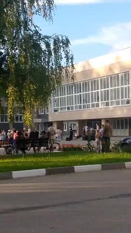 The video shows an outdoor event in front of a building with large windows. A group of people are gathered, some standing, some sitting. A band or musical group appears to be performing on a makeshift stage or raised area. There are instruments visible, and people are facing the performers. There are also trees and greenery in the foreground, suggesting a park or public space. The overall atmosphere seems relaxed and informal.