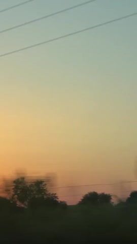 The video shows a blurred landscape with a sunset in the background. Power lines are visible against the sky. There are trees and a hint of hills on the horizon. The view appears to be from a moving vehicle.