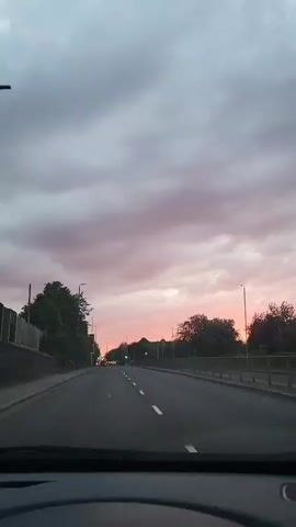 The view from inside a car shows a straight, two-lane road stretching into the distance under a sky tinged with a pink sunset.  Trees line both sides of the road, punctuated by streetlights and road signs.  The road markings are clear, indicating the lanes, and traffic lights ahead are green. The dashboard of the car is visible at the bottom of the frame.