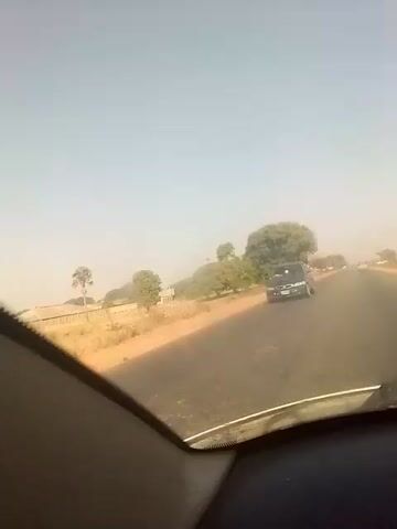 The video shows a view from inside a car driving on a paved road. Another car passes by on the left. The surrounding landscape appears dry, with sparse trees and sandy soil visible on the roadside. The sky is a light blue, and the overall visibility seems somewhat hazy. Music is playing in the background.