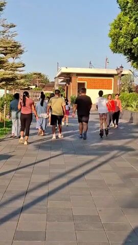 A group of people are walking on a paved path outdoors. The path is made of large, dark gray square tiles. On the left side of the path, there are trees with sparse, green foliage and some brown mulch. On the right side of the path, there are bushes and trees. In the background, there is a building with a white roof and brown brick walls. The sky is clear and blue. The sun is casting long shadows on the path. The people are walking away from the camera. Some are wearing casual clothing, while others are wearing athletic wear. One person is pushing a yellow cart with a black bin. The video is shot from a slight angle from behind the group.