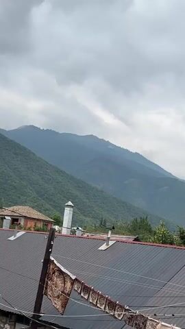 The video shows a view of mountains covered in dense green trees under a cloudy sky. In the foreground, there are several rooftops, some made of dark metal and others of reddish-brown tiles. There are also power lines and utility poles visible. A few buildings, including a house with an orange-tiled roof and a white structure that might be a chimney or minaret, are situated between the rooftops and the mountains. A string of rugs or tapestries hangs on a clothesline in front of the dark metal roof. The overall scene suggests a rural or semi-rural mountain village on an overcast day. There are no people visible in the video.