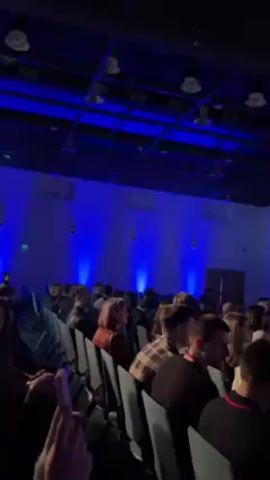 The video depicts a conference hall filled with attendees. They are seated in rows of chairs facing a large screen displaying a QR code and Russian text. The room is lit with blue spotlights, and several ceiling lights are visible. People appear to be mostly still, listening or watching the screen. Some individuals are partially obscured by those in front of them.