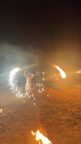 Two people are performing a fire dance on a sandy area at night. There are several fires visible, including a large one in the foreground and smaller ones associated with the dancers' props. The dancers are holding flaming objects that are emitting sparks. One dancer is wearing a light-colored dress, and the other is wearing a darker outfit. In the background, there are blurred figures of spectators and string lights. The sky is dark with some faint lights that could be stars or distant lights. The overall atmosphere appears to be festive or celebratory.