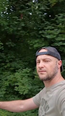 A man with short brown hair, a beard, and a black baseball cap is in the video. He is wearing a green t-shirt. The video appears to be taken outdoors in a natural setting with trees and greenery. The man is looking at the camera and smiling slightly. The video is shaky, suggesting it might have been recorded while the person was walking or moving. The background shifts from a dense green foliage to a path with trees and a cloudy sky.