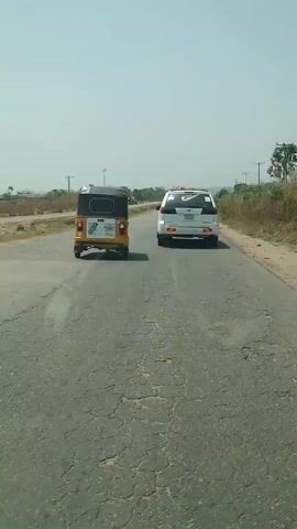 The video shows the rear view of two vehicles traveling on a cracked asphalt road. The vehicle in the foreground is a white SUV. In front of the SUV is a yellow and black three-wheeled vehicle, also known as a tuk-tuk. The sky is clear and bright. There are power lines along the side of the road, and some vegetation on the shoulder. In the distance, there are some hills. The camera is likely mounted inside another vehicle, following the two vehicles ahead.