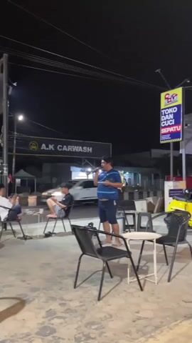 A man is standing and holding a microphone, possibly singing or speaking. He is wearing a blue t-shirt and navy blue shorts. Several people are seated at small tables in front of him. In the background, there is a sign that says "A.K CARWASH" and another sign with the text "TOKO DAN CUCI SEPATU." The scene appears to be an outdoor cafe or seating area at night, illuminated by artificial lights.