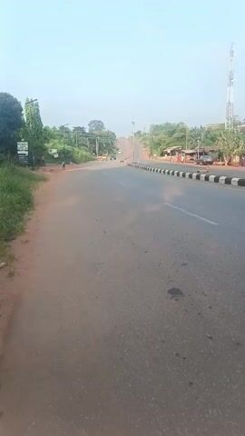 The video shows a paved two-lane road in what appears to be a rural or suburban area. The road has a slight uphill incline and black and white striped curb markers. There are some trees and greenery on the sides of the road, and buildings are seen further off in the distance. Several cars are driving on the road, and people are seen walking and standing near the roadside. There is a tall communication tower visible in the background to the right. The sky is clear and light blue. The video seems to be shot from the side of the road.