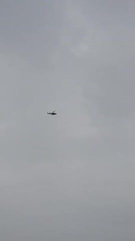 A helicopter flies across a cloudy sky. The helicopter is dark and silhouetted against the light gray clouds. Its rotors are spinning. The helicopter appears to be moving from left to right in the frame.