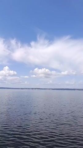 A video captures a view of a large body of water, likely a lake or sea, under a bright blue sky with scattered white clouds. The water is calm with gentle ripples. In the distance, several small, tree-covered islands are visible. The perspective suggests the video is being filmed from a moving boat, as evidenced by the slight disturbance and foam trailing in the lower left corner of the frame. The scene is peaceful and natural, with no people or man-made structures other than the implied boat, clearly visible. The time of day appears to be daytime, likely mid-morning or afternoon, given the lighting and clear sky.