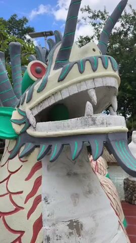 The video shows a close-up of a dragon statue's head, with its mouth open and sharp teeth visible. The dragon is colorful, with green accents and red markings on its scales. In the background, there are other statues, including a white statue of a seated Buddha and a larger white statue of a standing figure with a halo. There are also trees, plants, and a blue sky with clouds. The scene appears to be outdoors, possibly in a temple or garden. The camera is focused on the dragon statue, with the background slightly blurred. The video is short, lasting only a few seconds.