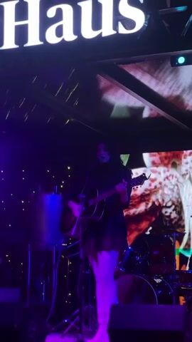 A woman with dark hair is singing and playing an acoustic guitar on a stage with a microphone. She is dressed in black and wearing a short skirt. Behind her is a drum set and a large screen displaying a blurred image. The stage is lit with purple and blue lights, and there is a sign reading "Haus BEER & MUSIC" in the background. The scene appears to be a live music performance at a bar or club.