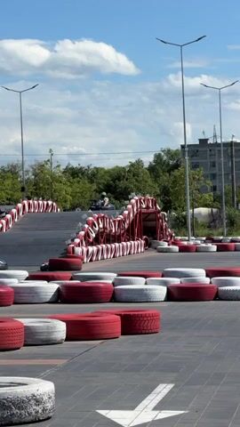 The video shows a go-kart race track on a sunny day with a blue sky and scattered clouds. The track is lined with red and white tires, acting as barriers. Several go-karts are visible on the track, some with drivers wearing helmets. In the background, there are trees, a tall building, and streetlights. One go-kart is seen going up a ramp. The camera pans slightly across the track.