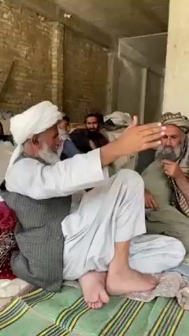 The video depicts four men seated on a patterned rug in what appears to be a partially constructed or unfinished building. The man in the foreground wears a white turban, light-colored clothing, and a dark vest; he sits cross-legged, leaning slightly back, with bare feet and crossed legs. To his right, another man, also bearded, wears a turban, a light-colored robe, and a patterned scarf; he has his hand to his chin, seemingly in contemplation. Behind them, a third man with a beard sits with his arms crossed, and in the background, a fourth man looks towards the camera. The environment includes brick walls and concrete pillars, with bags of material visible in the background. The overall impression is of a casual gathering in a somewhat rustic or unfinished setting.