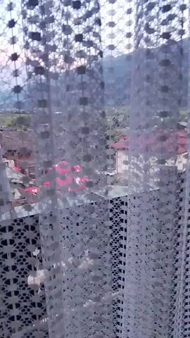 A video shot from behind a white lace curtain shows a view of a village nestled in a valley with mountains in the background. The village appears to be made up of traditional houses with red-tiled roofs. There is an outdoor seating area with red umbrellas and tables, suggesting a cafe or restaurant. The sun is low in the sky, casting a soft light over the scene. The video pans slowly from left to right, revealing more of the village and the surrounding landscape.