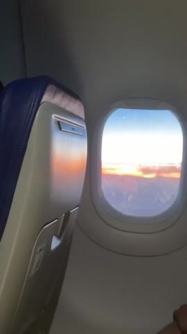The view from an airplane window showcases a vibrant sunset. The seat armrest, reflecting the orange and red hues, is on the left side of the frame. Outside the window, a clear blue sky meets a thick layer of clouds illuminated by the setting sun. The landscape below appears dark and indistinct.