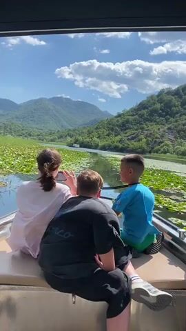 The video shows three individuals, two children and one adult, from behind, seated on a boat. They are looking out at a scenic landscape. The person in the center, appearing to be an adult, is wearing a dark t-shirt and black pants. To their left is a person, likely a female, wearing a light pink shirt and gray pants, holding a phone and taking a picture. To the right is a young boy in a blue t-shirt and green shorts. They are on a boat that is moving through a body of water covered with lily pads. In the background, there are lush green mountains under a bright blue sky with fluffy white clouds. The scene appears to be a serene nature tour.