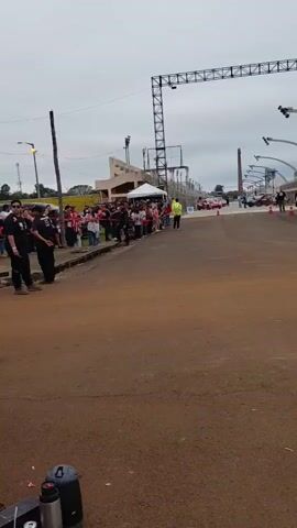 A group of spectators stands behind a barrier, watching a rally car event. The road surface is dirt and some dust is kicked up. A fire truck is parked on the right side of the frame. A few rally cars are visible in the distance, along with some cones marking the course. The sky is overcast.