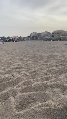 The video displays a ground-level view of a sandy beach. The sand is textured with small ripples, indicating it has been disturbed. In the background, a train is traveling along tracks that run parallel to the beach. Beyond the train tracks, there are buildings that appear to be a town or city. Further in the background, there are a few people visible on the beach, some sitting under beach umbrellas. The sky is overcast.