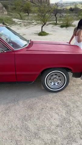A red classic convertible car, possibly a Chevrolet Impala, is parked on a dirt path. The car features a matte red exterior, chrome accents, and distinct red and white striped seats. The video begins with a side view of the car, showing its front wheel and door, then slowly pans to the back, revealing the rear wheel and trunk. In the background, unique geological formations known as "fairy chimneys" rise from the landscape, accompanied by scattered green vegetation and trees. Dirt paths wind through the hills behind the car. The scene is set outdoors during daylight hours in Çavuşin Köyü, Turkey, showcasing the distinctive natural beauty of Cappadocia alongside the vintage vehicle.