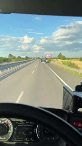A first-person perspective video shows a view from the driver's seat of a truck driving on a highway. The road is asphalt with white lane markings. In the distance, a blue and red bus is driving in the same direction. On the right side of the highway, there is a billboard with an advertisement for Milka chocolate. The sky is blue with scattered white clouds. The truck's dashboard displays various information, including speed and engine status.