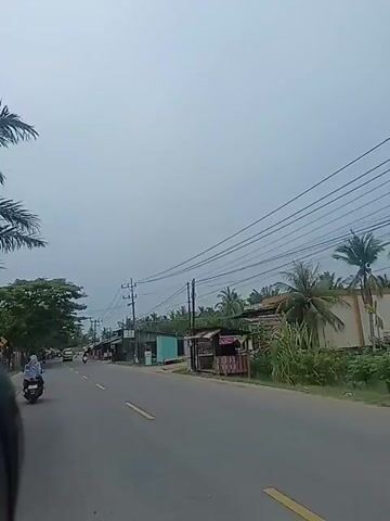 The video shows a view from a moving vehicle, likely a motorcycle, traveling on a road. The road is asphalt with yellow lane markings. On the left side of the road, there are trees and a few buildings. Several motorcycles are visible, with one in the foreground on the left and others further down the road. A yellow truck is also seen driving on the road. On the right side of the road, there are small shops or stalls with corrugated metal roofs. Power lines stretch across the sky, supported by utility poles. The sky is overcast. The scene appears to be a rural or suburban area.